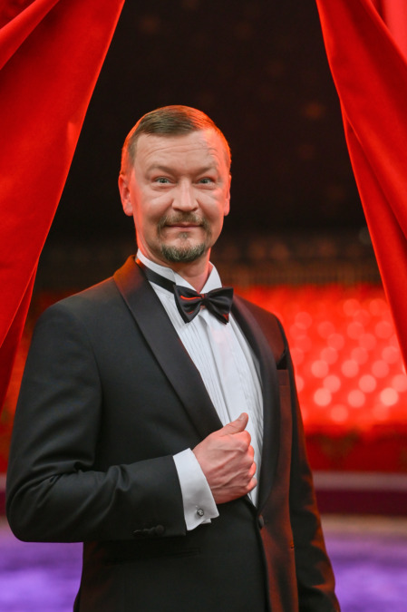 Sirkusjohtaja Calle Jernström Jr seisoo frakki päällä punaisten esirippujen välissä, taustalla sirkusteltan punaiset valot