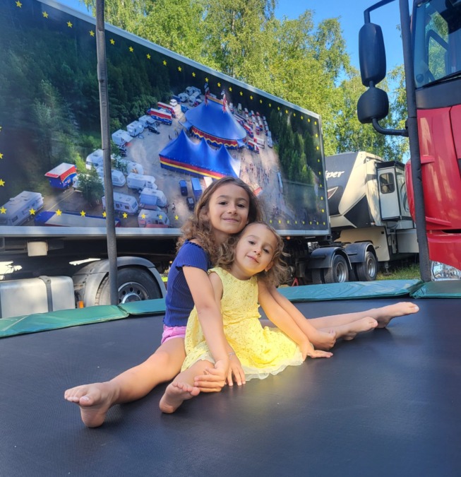Kaksi lasta istuu trampoliinilla sirkusvaunujen edessä, taustalla Sirkus Finlandian rekka, jonka kyljessä on kuva sirkusalueesta.
